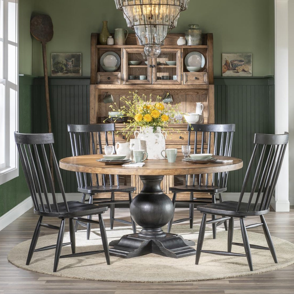 Rustic round dining table with four black chairs, set in front of a wooden hutch in a green room.