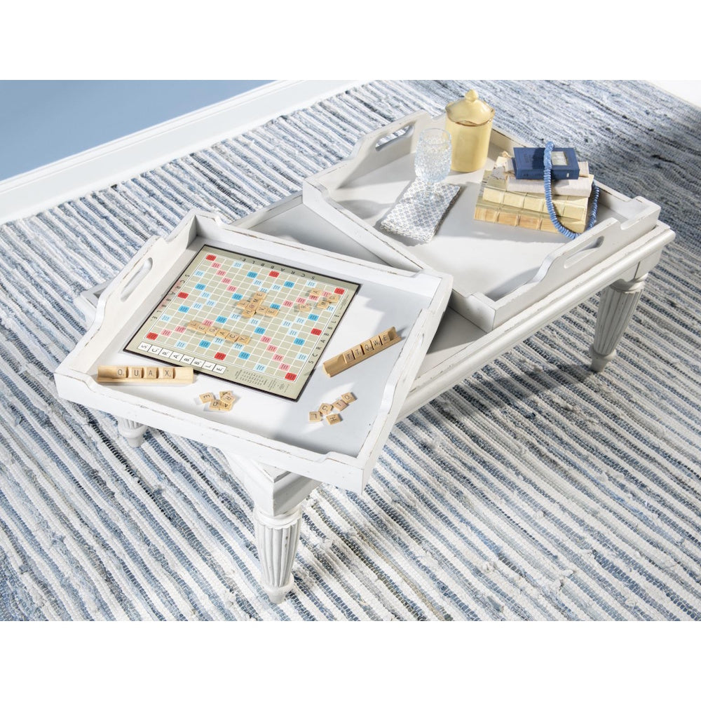 White cocktail table with removable tray tops holding a Scrabble game and decor, on a blue textured rug.