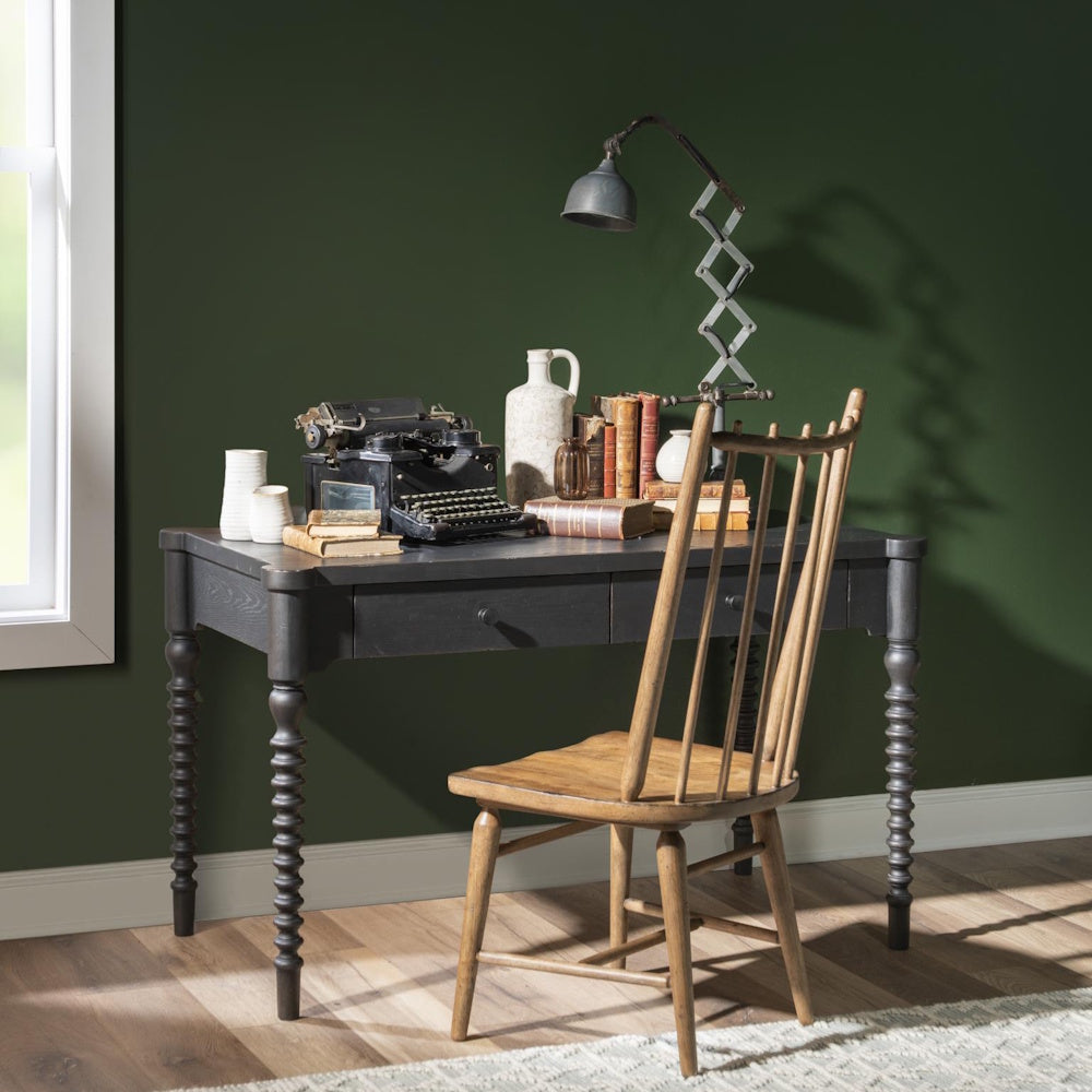 Vintage black bobbin-leg writing desk with a wooden chair, typewriter, and books against a green wall.