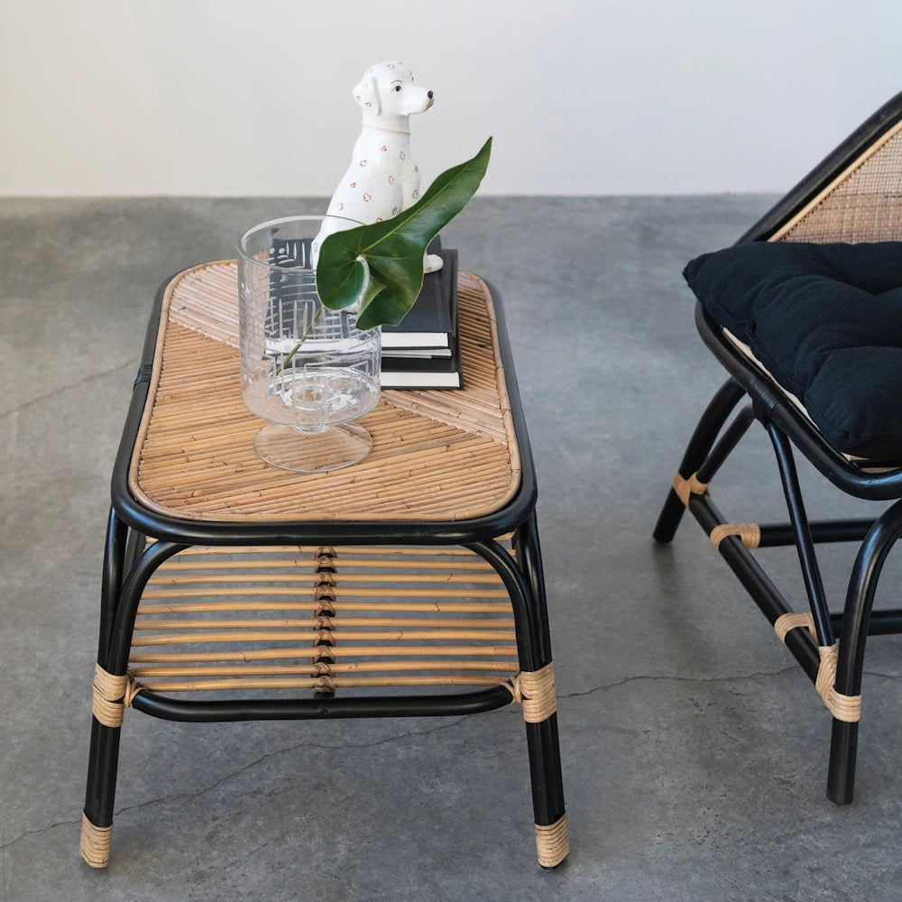 Wicker coffee table with glass vase, ceramic dog figurine, and monstera leaf beside a black rattan armchair with a cushion.