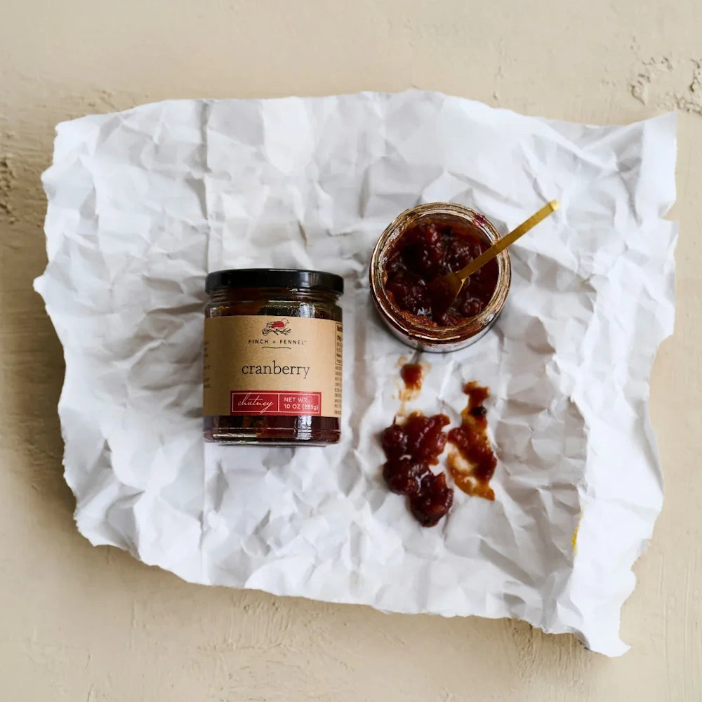 Jar of cranberry chutney beside an open jar and spoon on crumpled white paper.