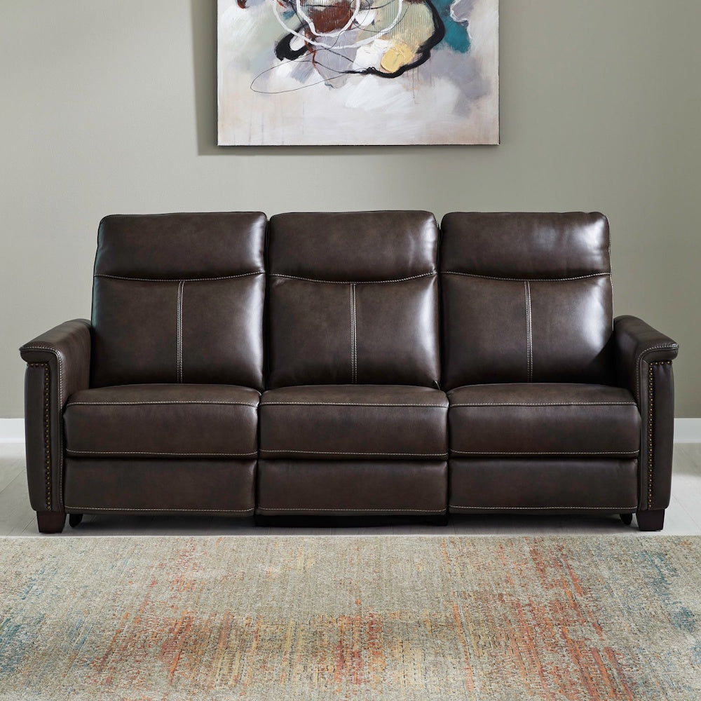 Modern dark brown faux leather reclining sofa with nailhead trim on a patterned area rug.