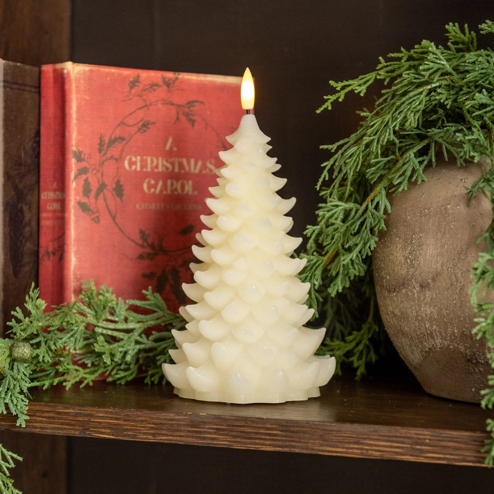 Lit cream tree-shaped flameless candle on a wooden shelf next to a vintage book and greenery.