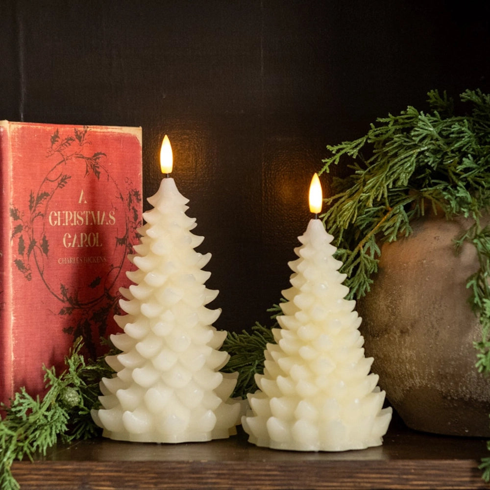 Two lit cream tree candles next to a red book and green foliage on a shelf.