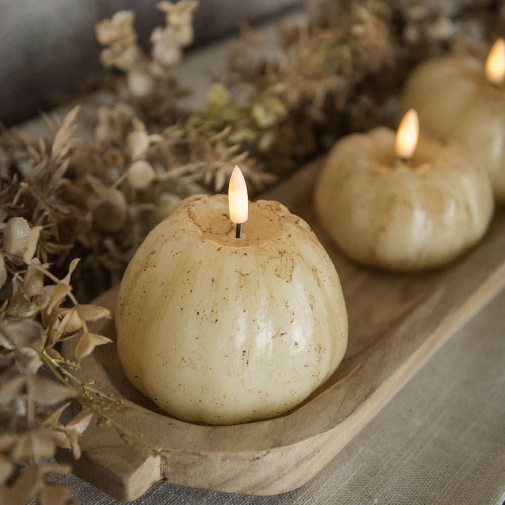 Close-up of speckled cream pumpkin 3D flame candles arranged in a narrow wooden dough tray with fall foliage.