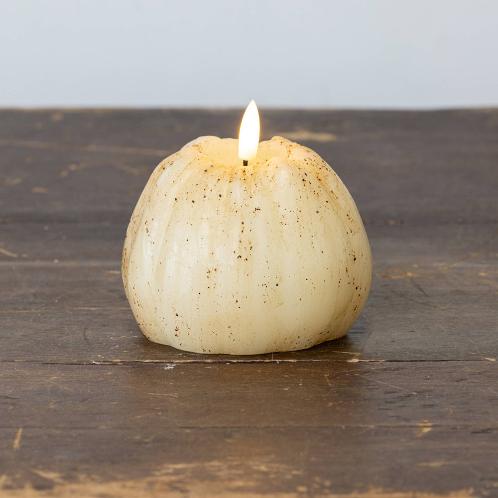 Lit cream 3D flame pumpkin candle with realistic dark speckles sitting on a rustic wooden table.