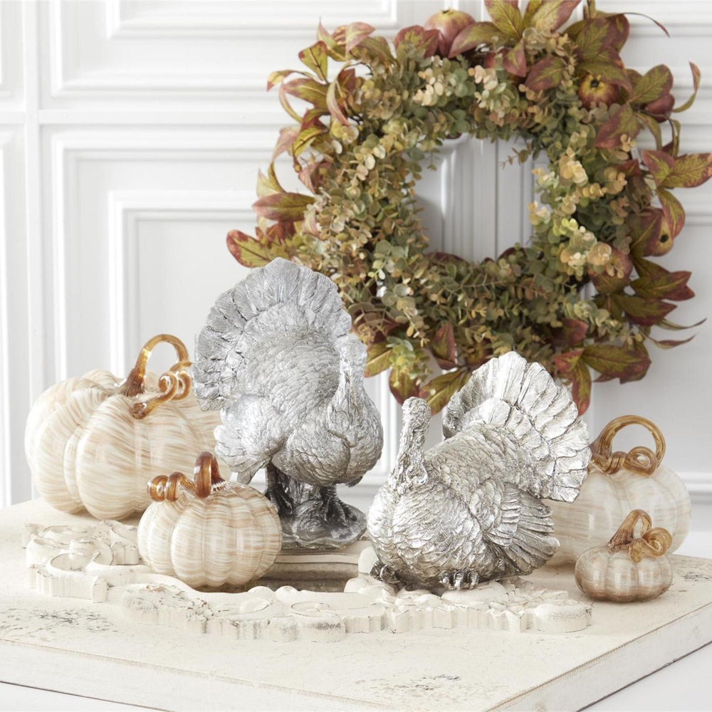 Silver turkey figures and cream glass pumpkins arranged in front of a fall wreath.