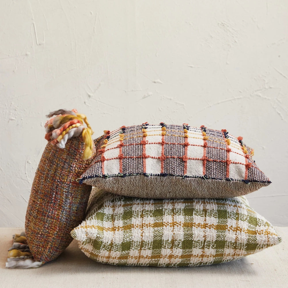 A stack of two plaid woven pillows next to a textured, fringed bolster pillow against a rustic wall.