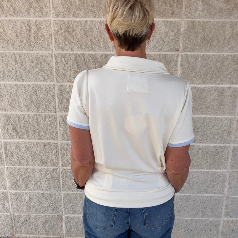 Back view of a woman in a cream short-sleeve knit top with light blue trim and a collar, standing against a brick wall.