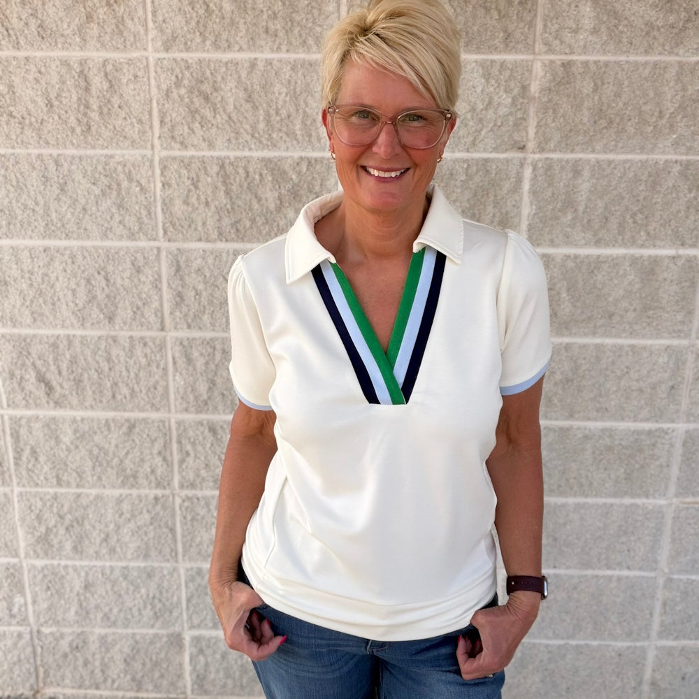 Smiling woman wearing a cream V-neck polo top with green and blue striped trim against a gray brick wall.