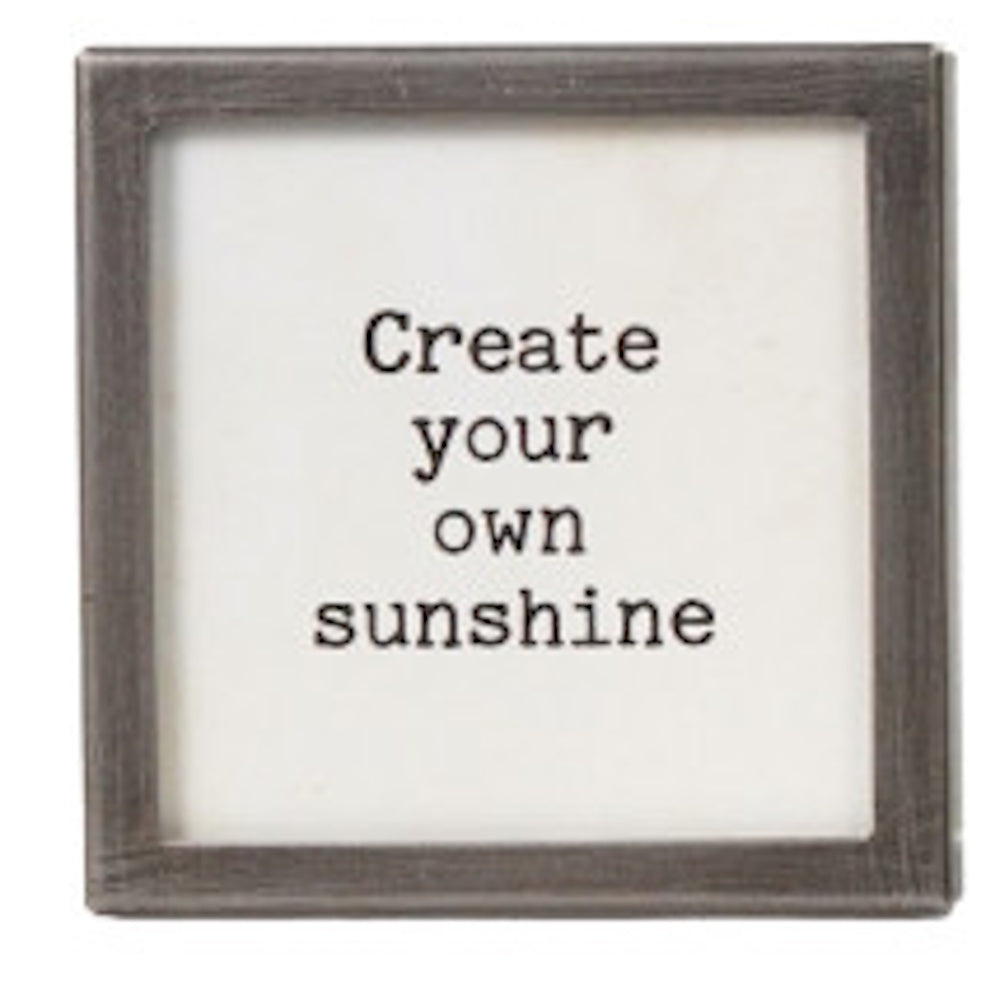 A square silver-framed sign with the text "Create your own sunshine" in black typewriter font.