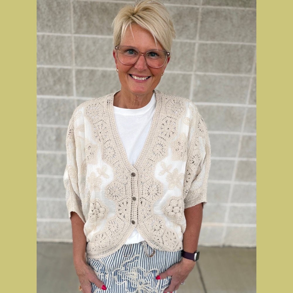 Smiling woman with short blonde hair wearing a beige crochet cardigan over a white top and striped pants.