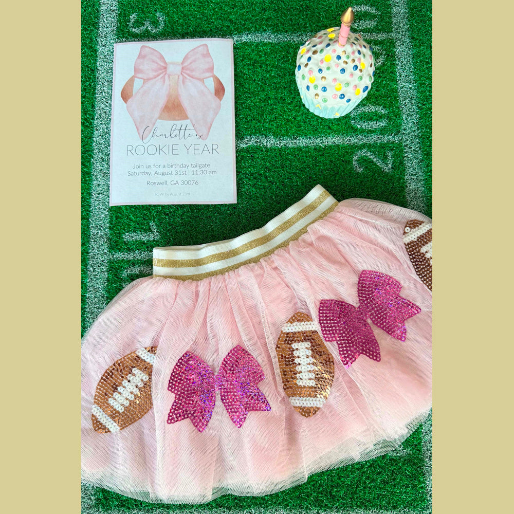 Pink tutu with sequin footballs and bows, a birthday invitation, and a ceramic cupcake on a green turf field.