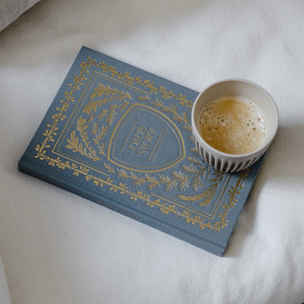 Blue "Dad's Story" keepsake journal with gold leaf patterns and a cup of coffee on a white textured surface.
