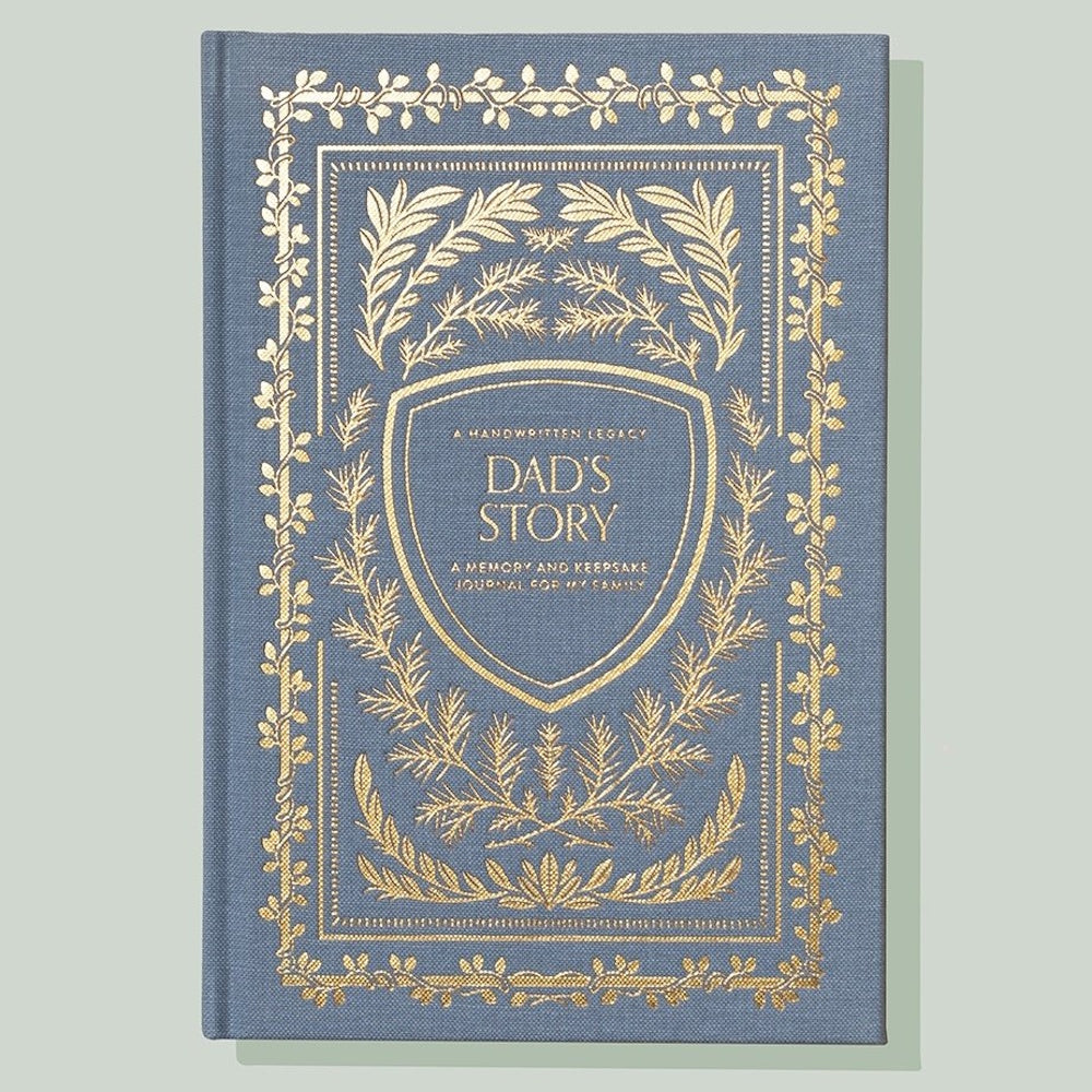 Blue fabric "Dad's Story" memory journal with gold foil leaf patterns and a central crest design on the cover.
