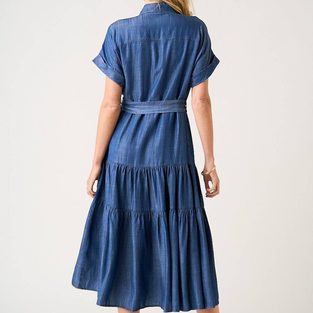 Back view of a woman wearing a dark denim washed Tencel chambray maxi shirt dress with a tiered skirt and belted waist.
