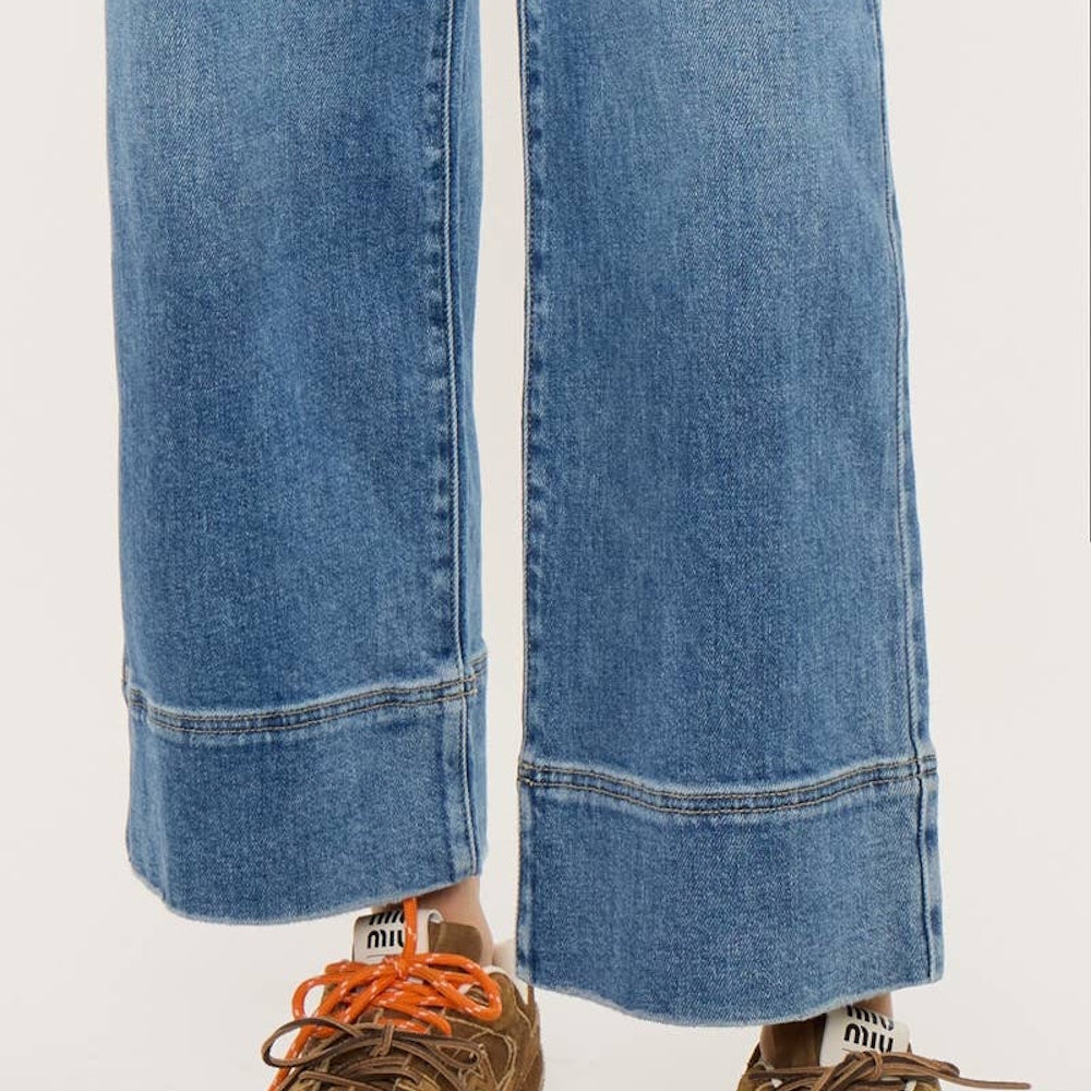 Close-up view of wide-leg blue denim jeans featuring wide hem stitching, paired with brown sneakers.