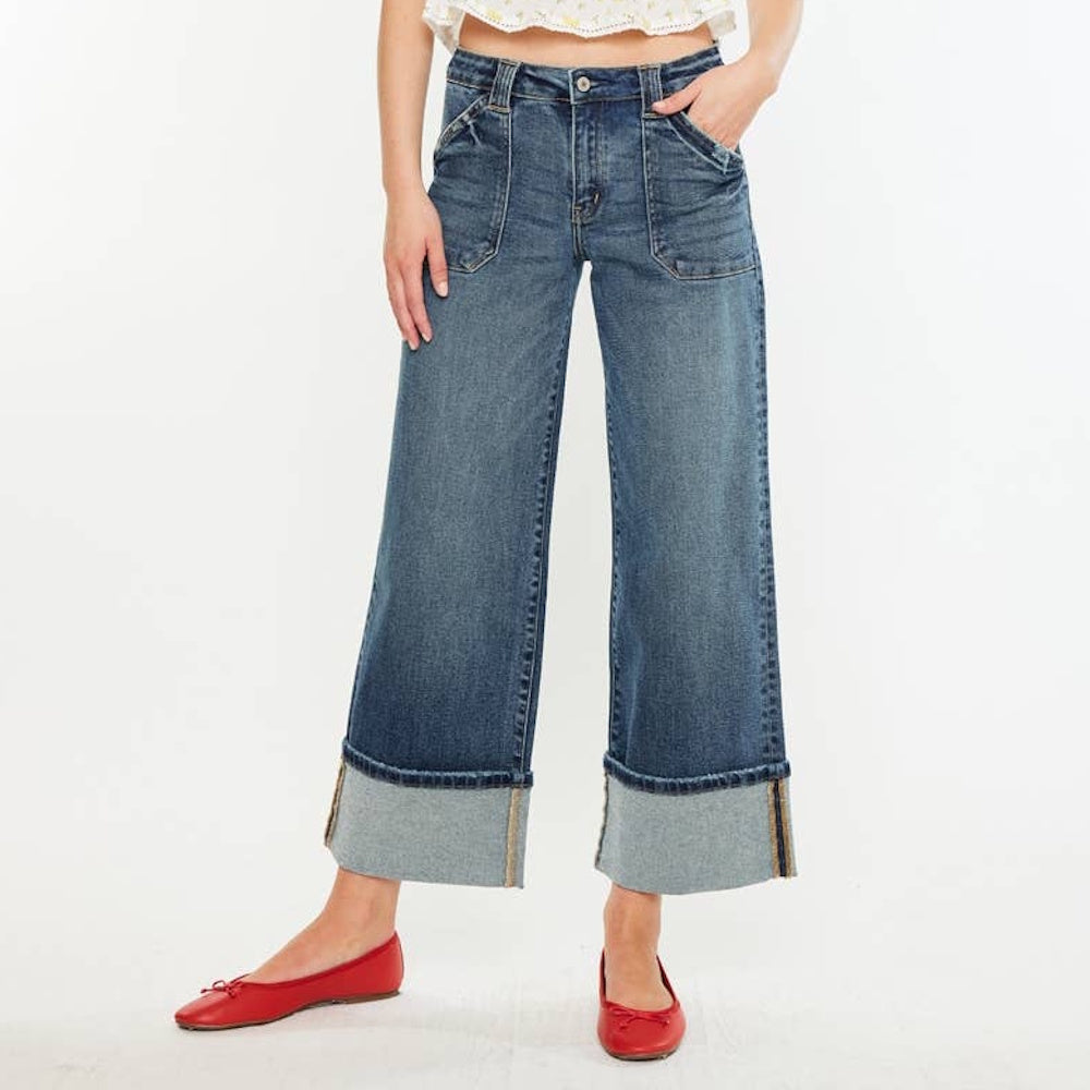 A woman wearing mid-rise wide-leg blue jeans with large cuffed hems, front utility pockets, and red flats.
