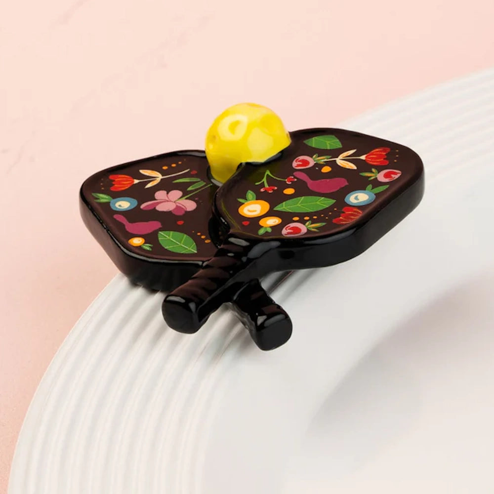 A decorative Nora Fleming mini featuring two black pickleball paddles with floral patterns and a yellow ball.