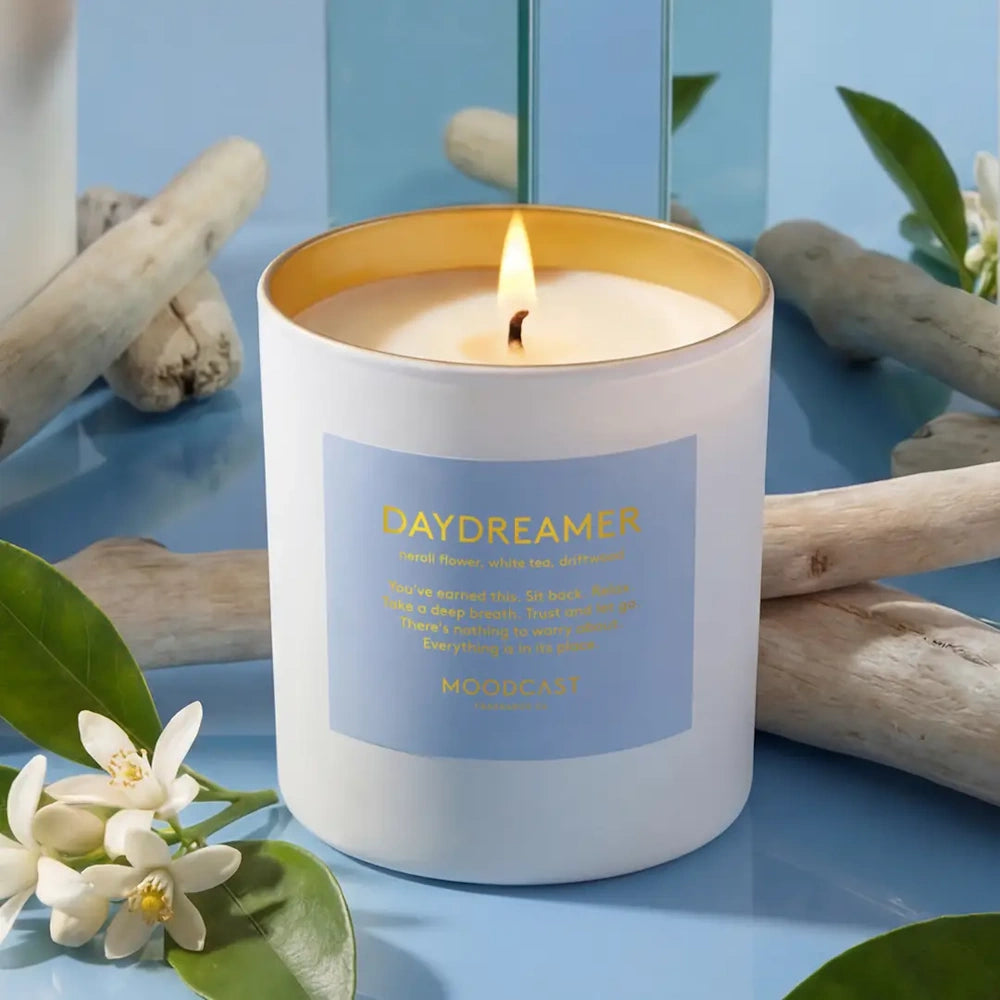 Lit Moodcast Daydreamer candle surrounded by white flowers, leaves, and pieces of driftwood.