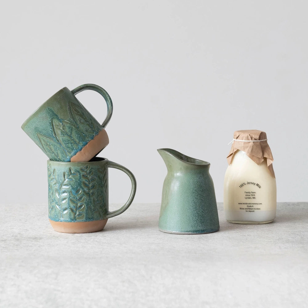 Handmade green ceramic mugs and creamer set next to a bottle of 100% Jersey Milk on a neutral tabletop.