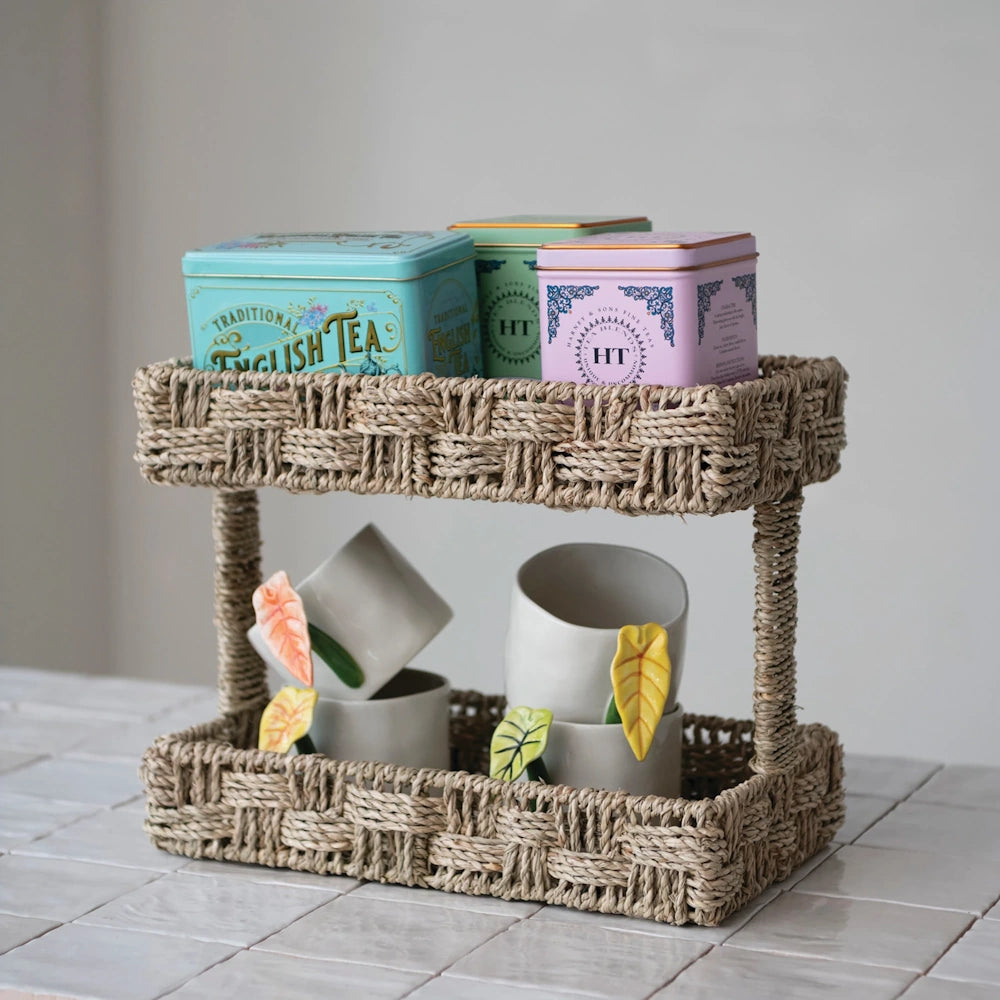Woven 2-tier seagrass tray styling tea tins on top and leaf-adorned ceramic cups below on a tiled counter.