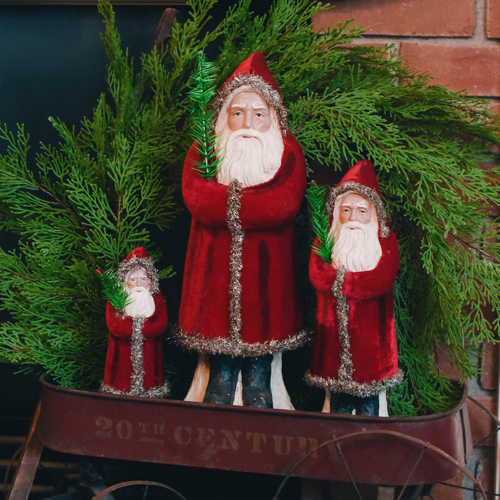 Three vintage Belsnickle Santa figures in deep red velvet coats with tinsel trim in a red metal wagon.