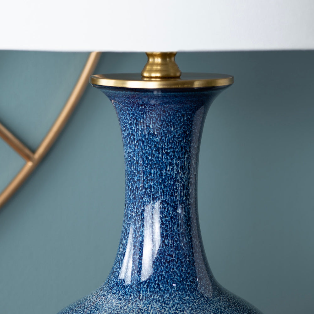 Close-up of a blue speckled ceramic lamp base with gold metal hardware and a white drum shade against a blue wall.