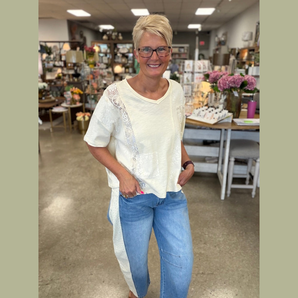 A woman in a cream lace-trimmed top and light-wash barrel jeans with side floral panels smiles in a shop.