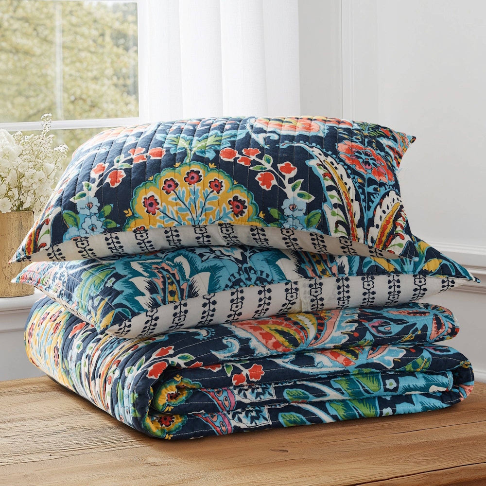 Stacked colorful paisley floral quilted pillow shams and quilt folded neatly on a wooden table.