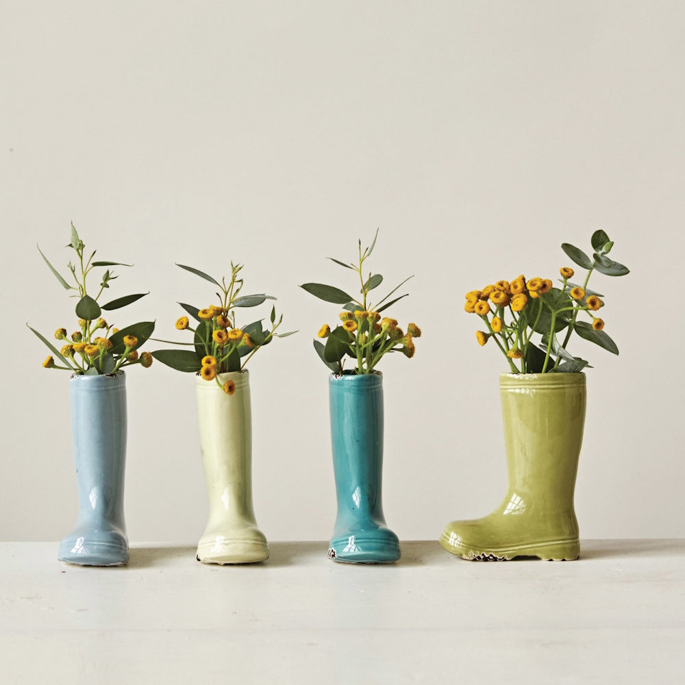 Four ceramic boot-shaped vases in pastel colors hold small yellow flowers and green leaves on a white surface.