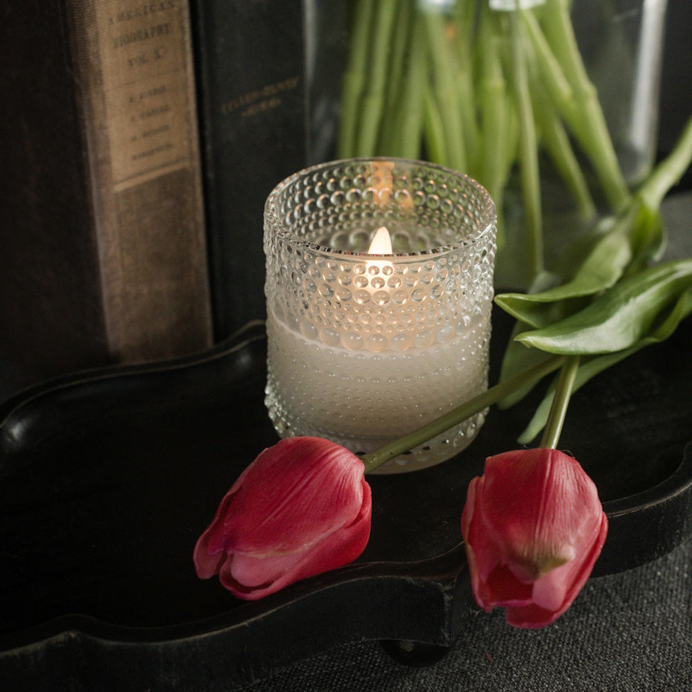 Clear dotted glass candle glowing on a tray next to pink tulips and antique books.
