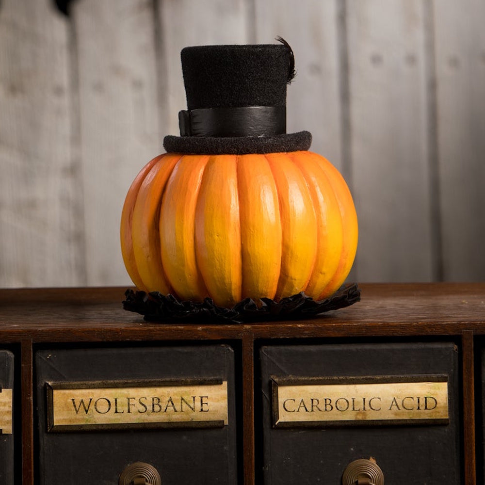 Back view of an orange pumpkin wearing a black top hat, sitting on vintage wooden drawers.