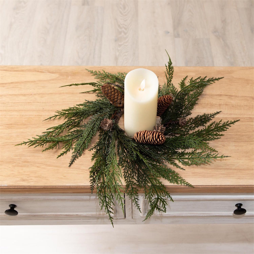 White pillar candle in a faux cedar pinecone ring wreath, placed on a wooden table.