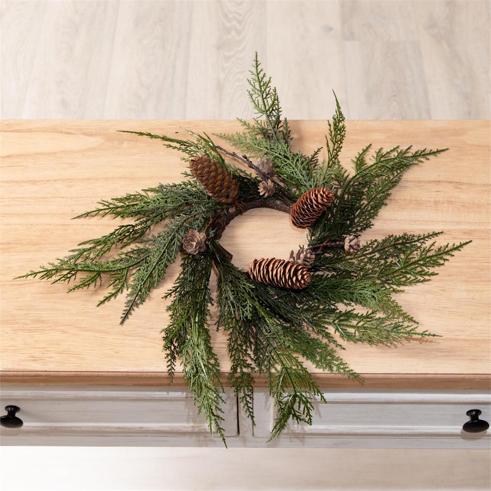 Faux cedar candle ring wreath with pinecones resting on a light wooden tabletop.