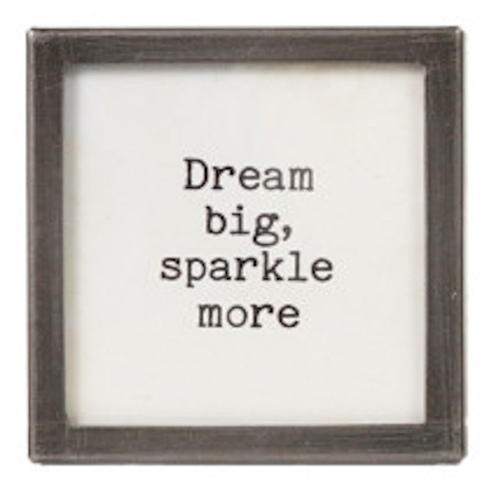 A square silver-framed sign with the text "Dream big, sparkle more" in black typewriter font.
