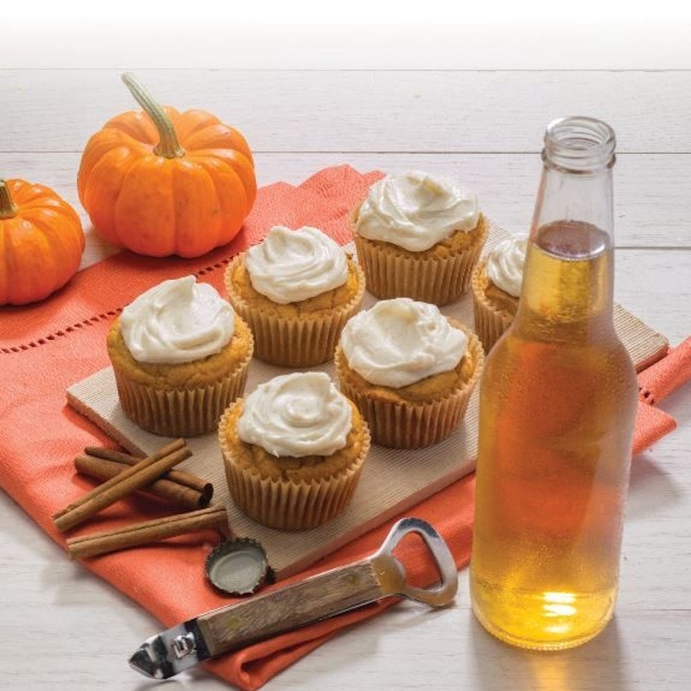 Frosted pumpkin cupcakes arranged next to a bottle of beer, cinnamon sticks, and mini pumpkins.