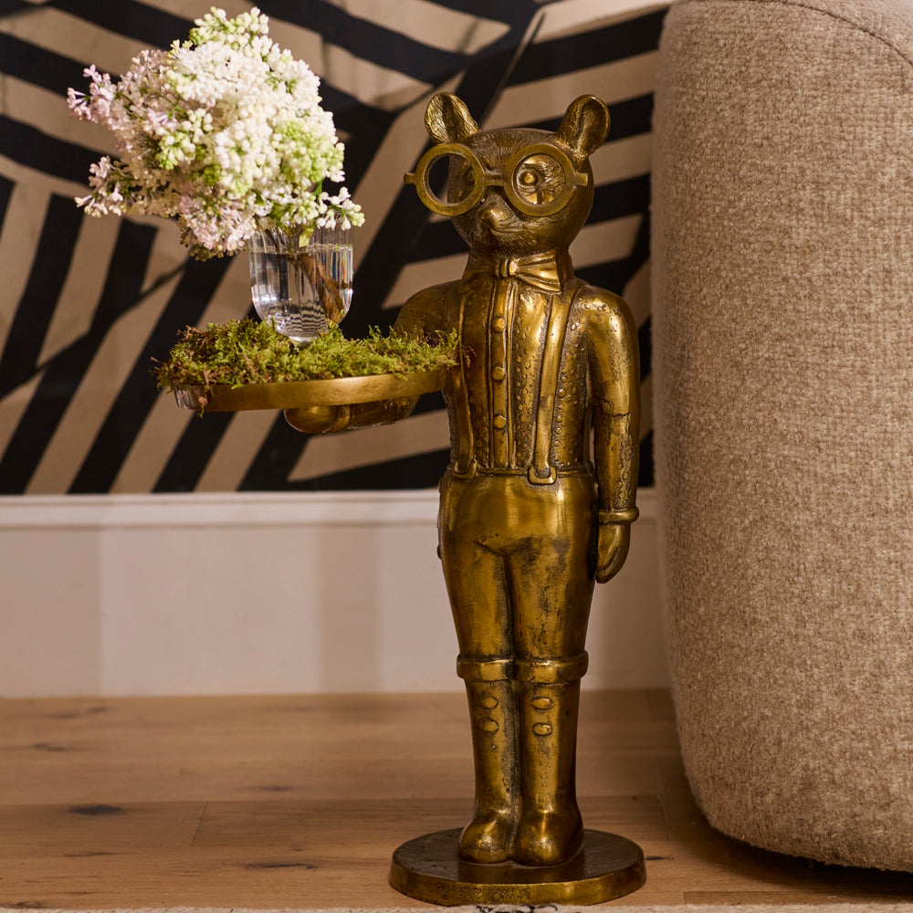 Brass butler statue wearing round glasses, holding a tray with white flowers next to a sofa.