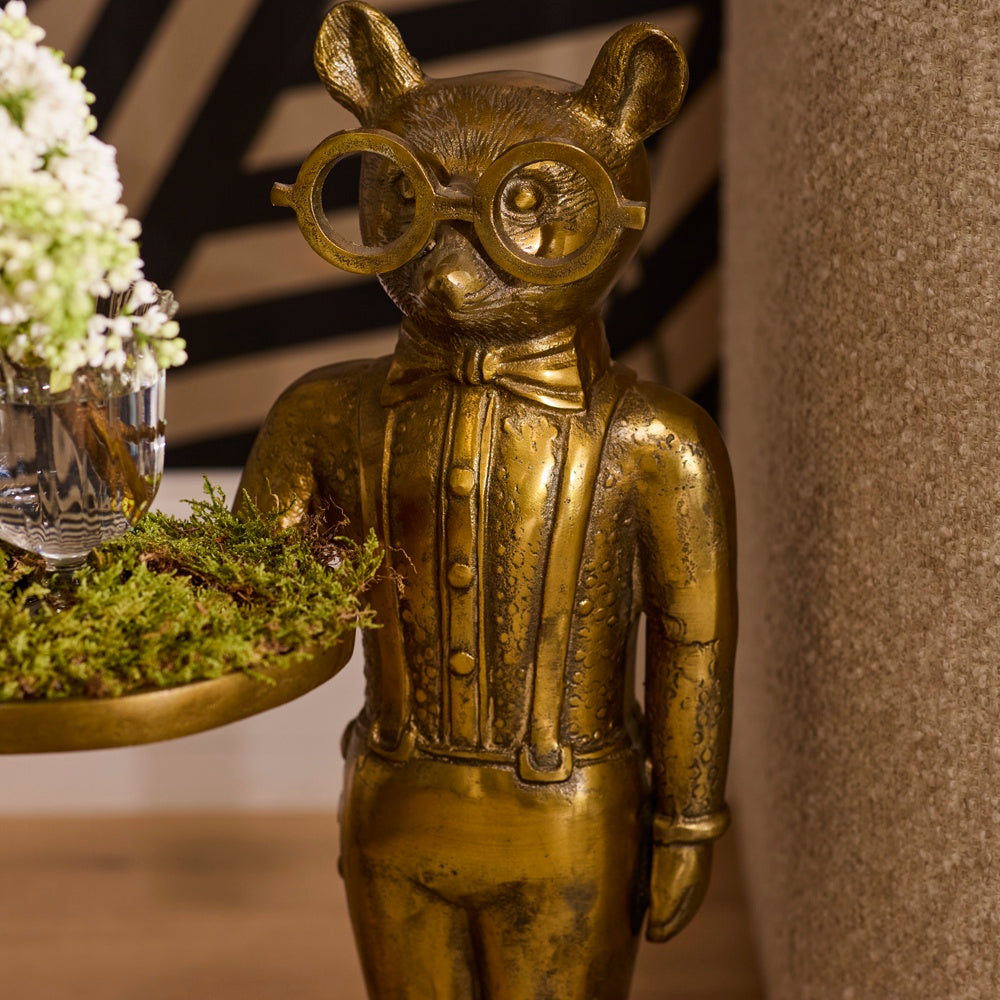 Close-up of a gold-finished bear butler statue wearing glasses, a bow tie, and suspenders, holding a tray with a small floral arrangement.