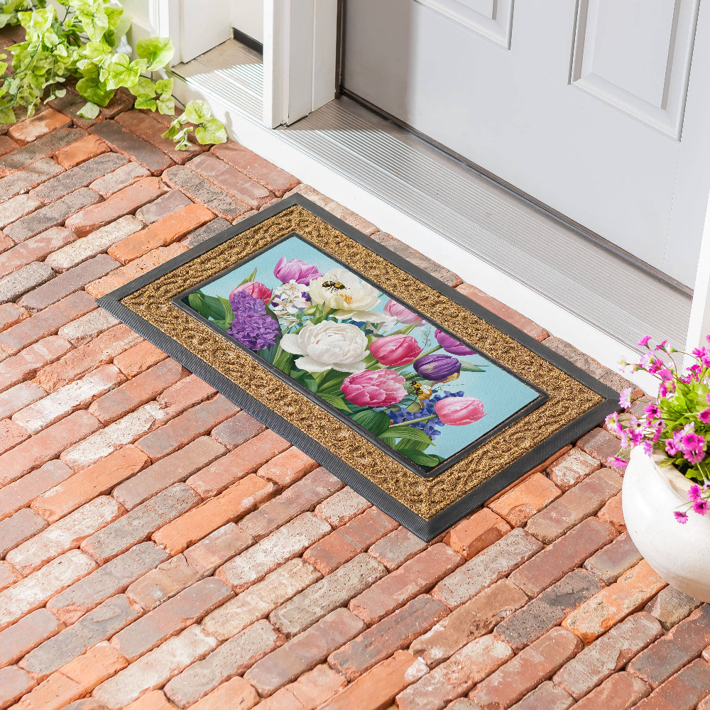 A decorative spring floral doormat with a coir border sits on a red brick entryway by a white front door.