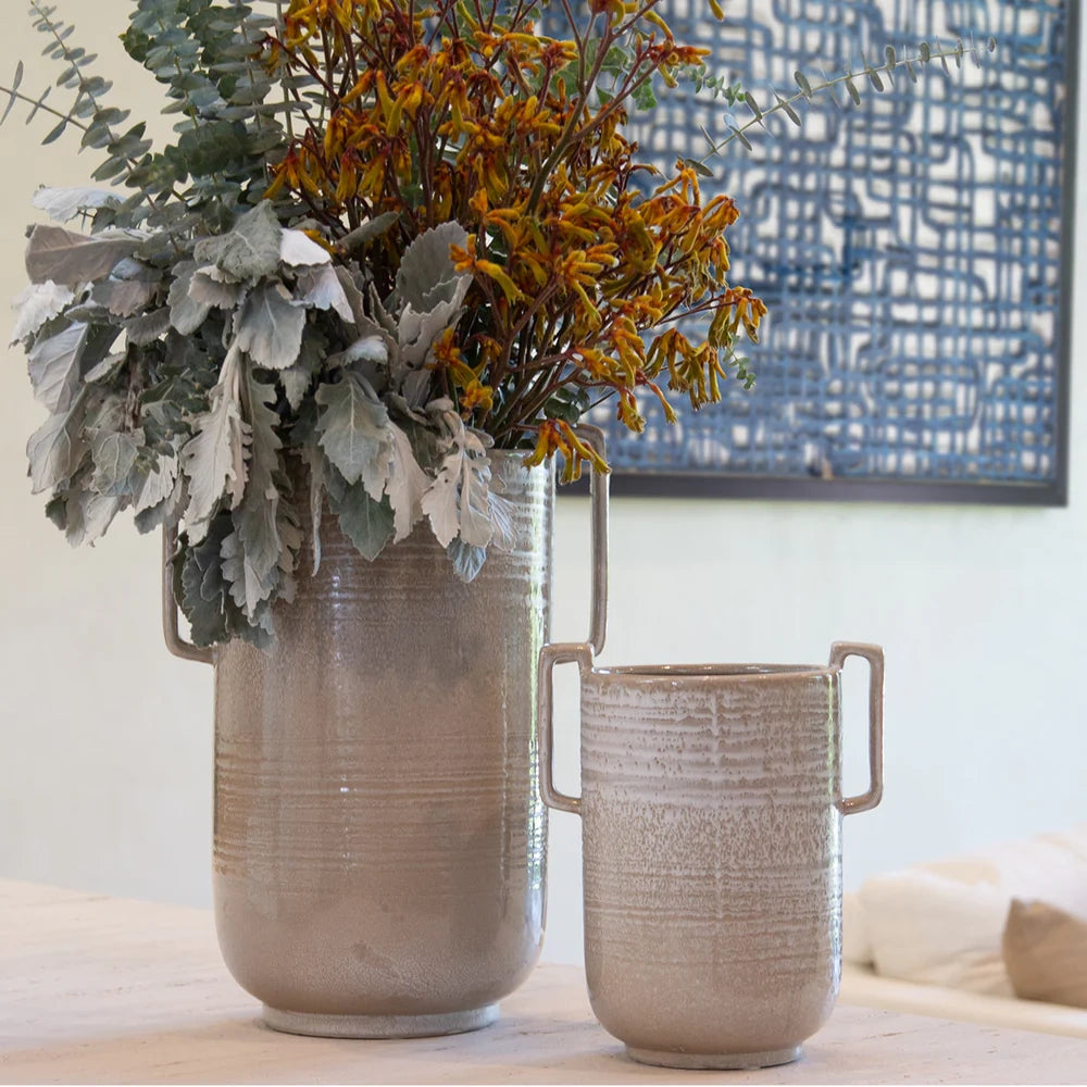Two textured ceramic vases with handles, one tall with yellow flowers and greenery, on a neutral tabletop.