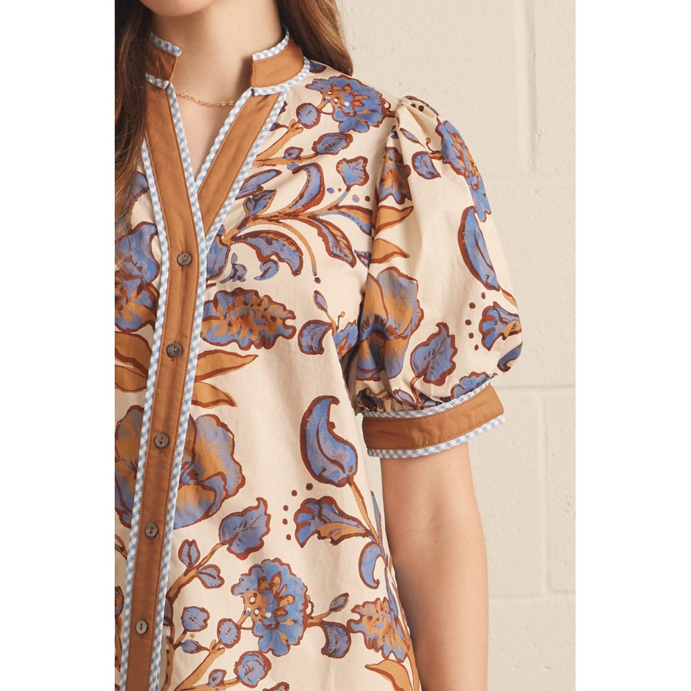 Close-up of an ecru floral print top featuring a brown button placket and blue checked piping.