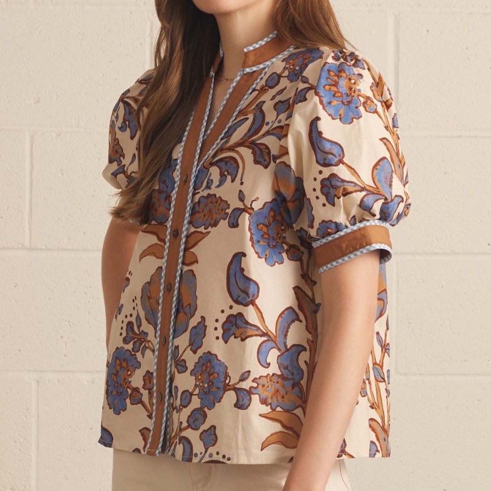 Three-quarter view of an ecru floral print top featuring a brown placket and blue checked piping.