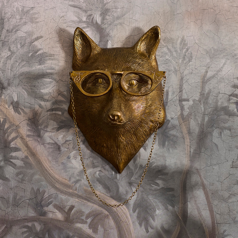 Brass fox head wall mount wearing cat-eye glasses with a decorative hanging chain on patterned wallpaper.
