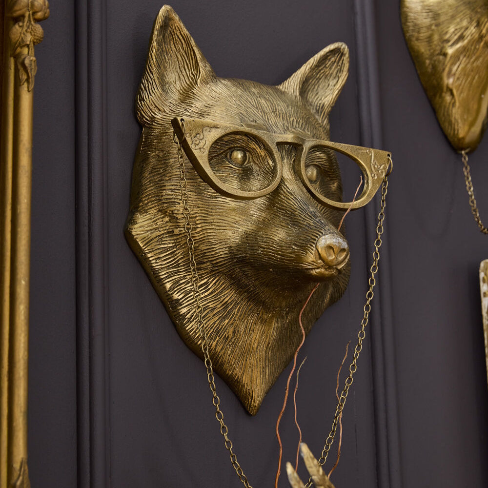 Gold fox head wall mount wearing cat-eye glasses with a hanging chain, displayed on a dark gray wall.