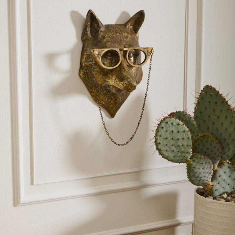 Bronze fox head wall mount wearing ornate glasses with a hanging chain, mounted next to a potted cactus.