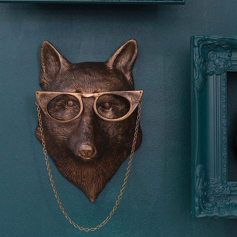 Bronze fox head wall mount wearing cat-eye glasses, mounted on a dark teal textured wall beside a frame.