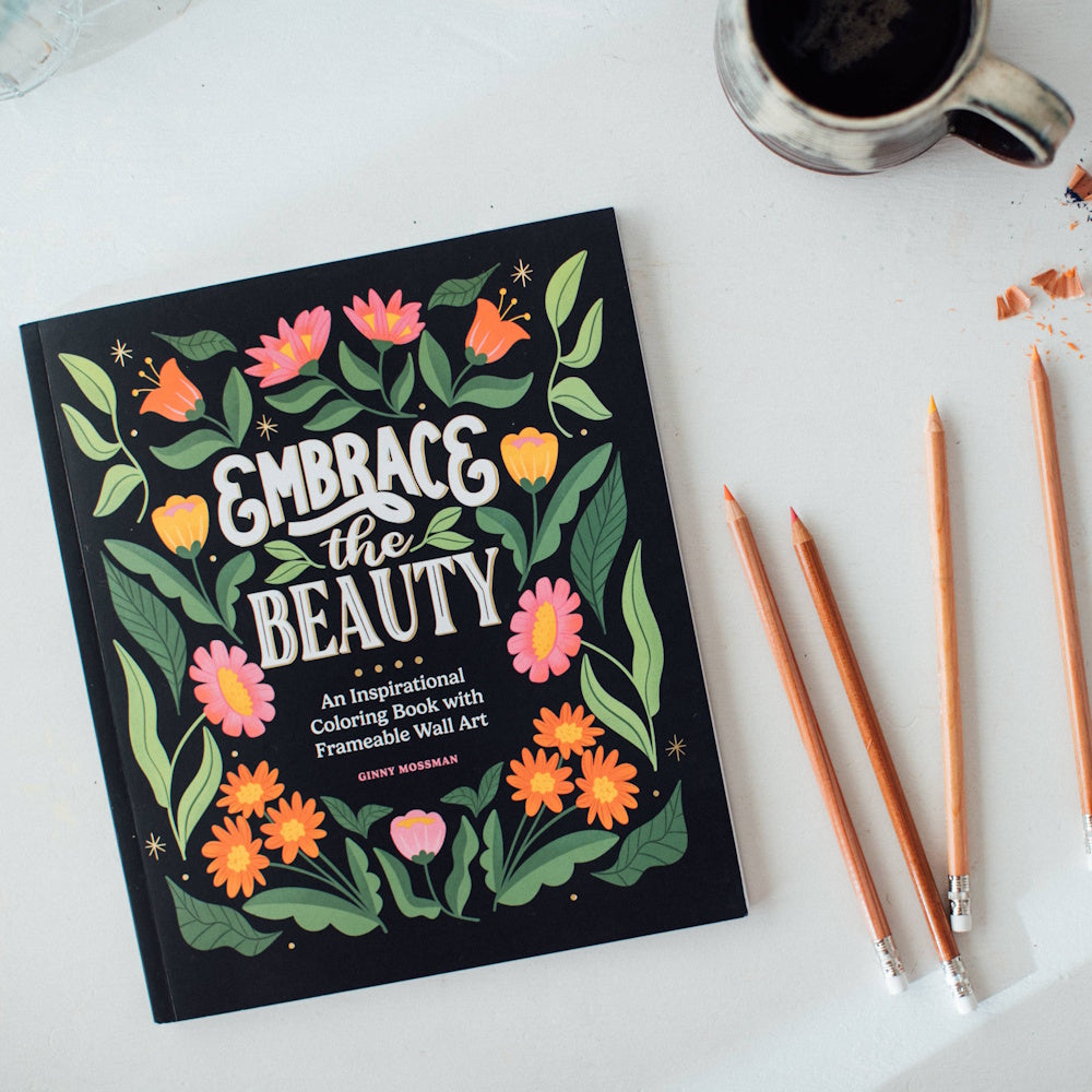 Embrace the Beauty inspirational coloring book by Ginny Mossman on a desk with colored pencils and a cup of coffee.