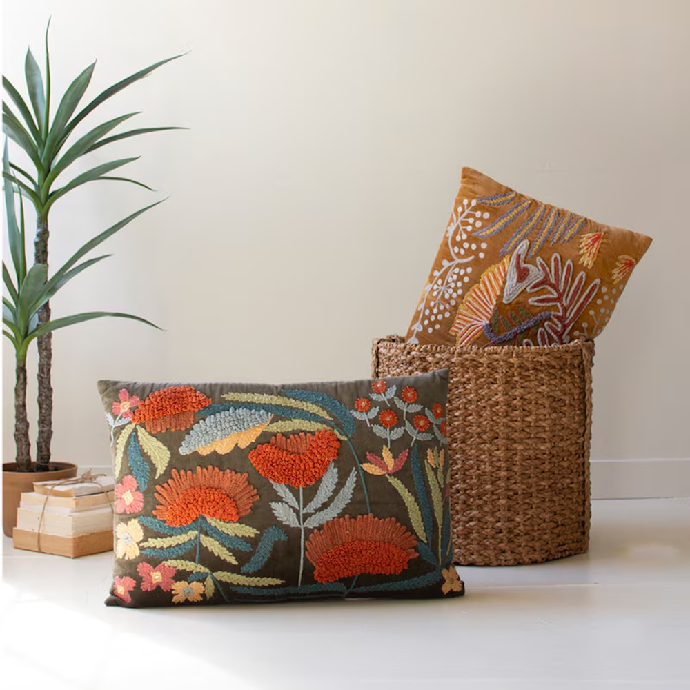 Embroidered throw pillows in brown and grey styled in a bright room with a plant and woven basket.