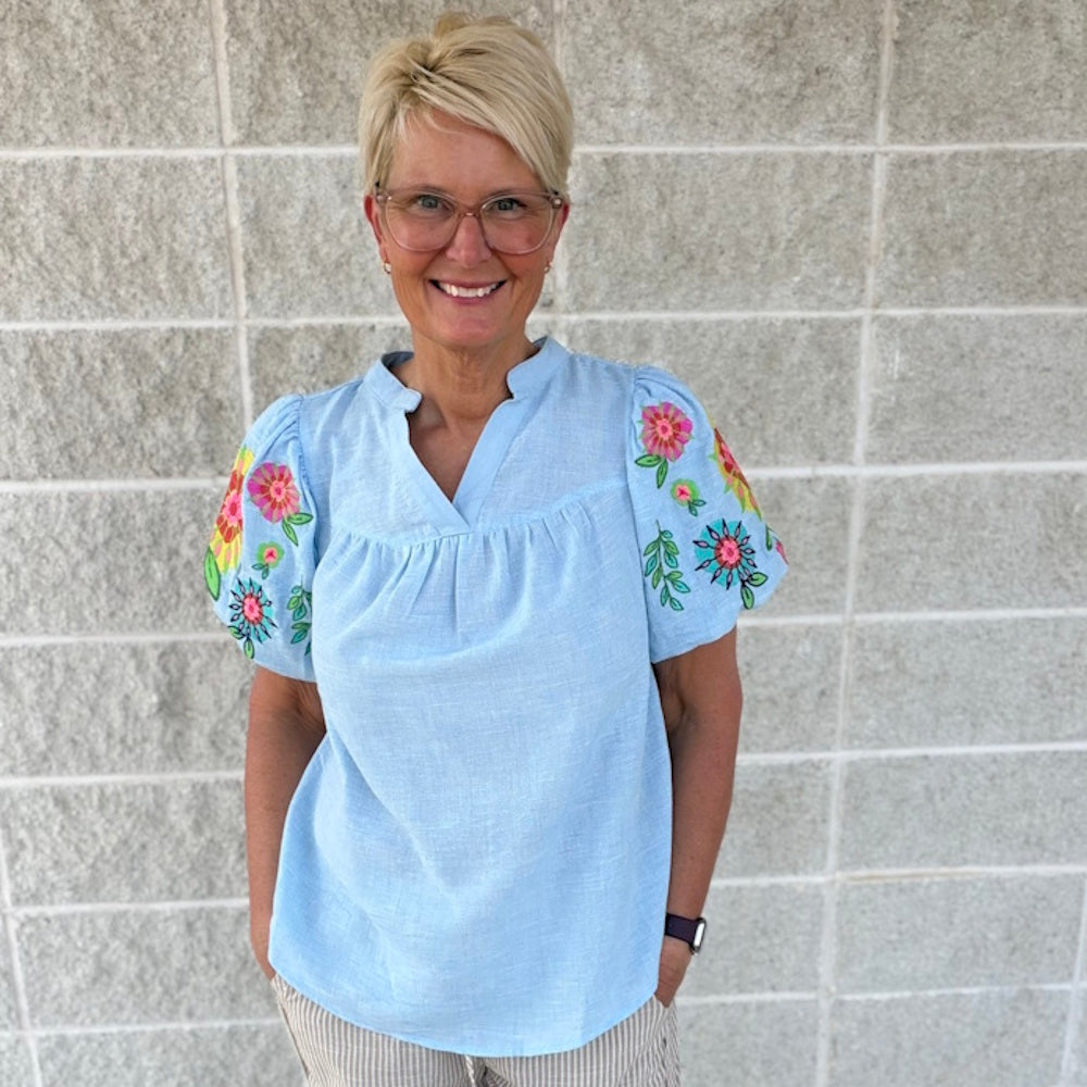 Woman wearing a light blue split-neck top with colorful floral embroidery on the puff sleeves.